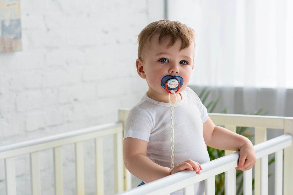 Baby  in white t-shirt standing in crib with pacifier in mouth